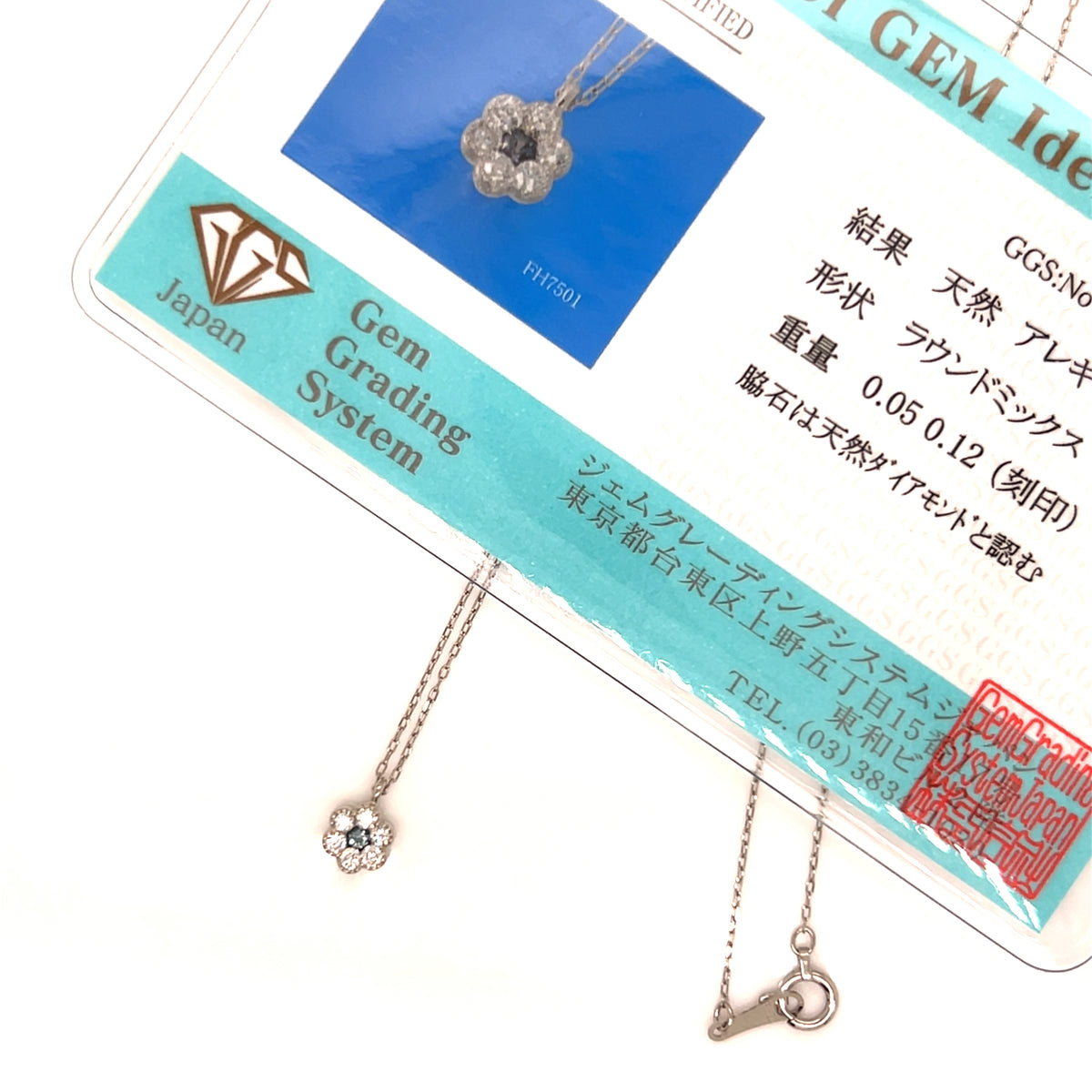 A close-up of a silver-toned flower-shaped pendant necklace with a clear center stone, hanging from its branded Gem Grading System display card. The card includes Japanese text and a blue inset showing the pendant.