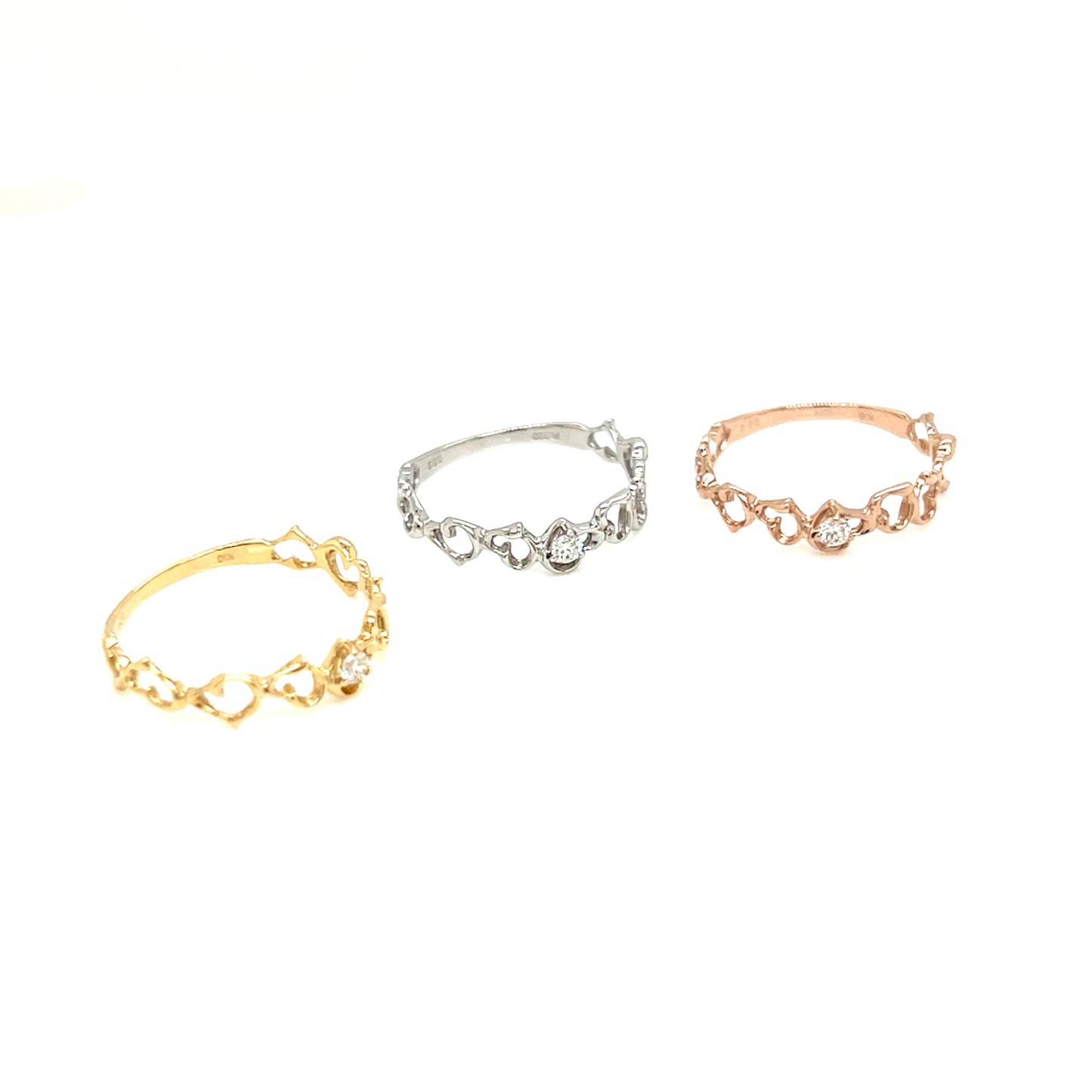 Three delicate heart-link rings in yellow gold, silver, and rose gold, arranged on a plain white background.