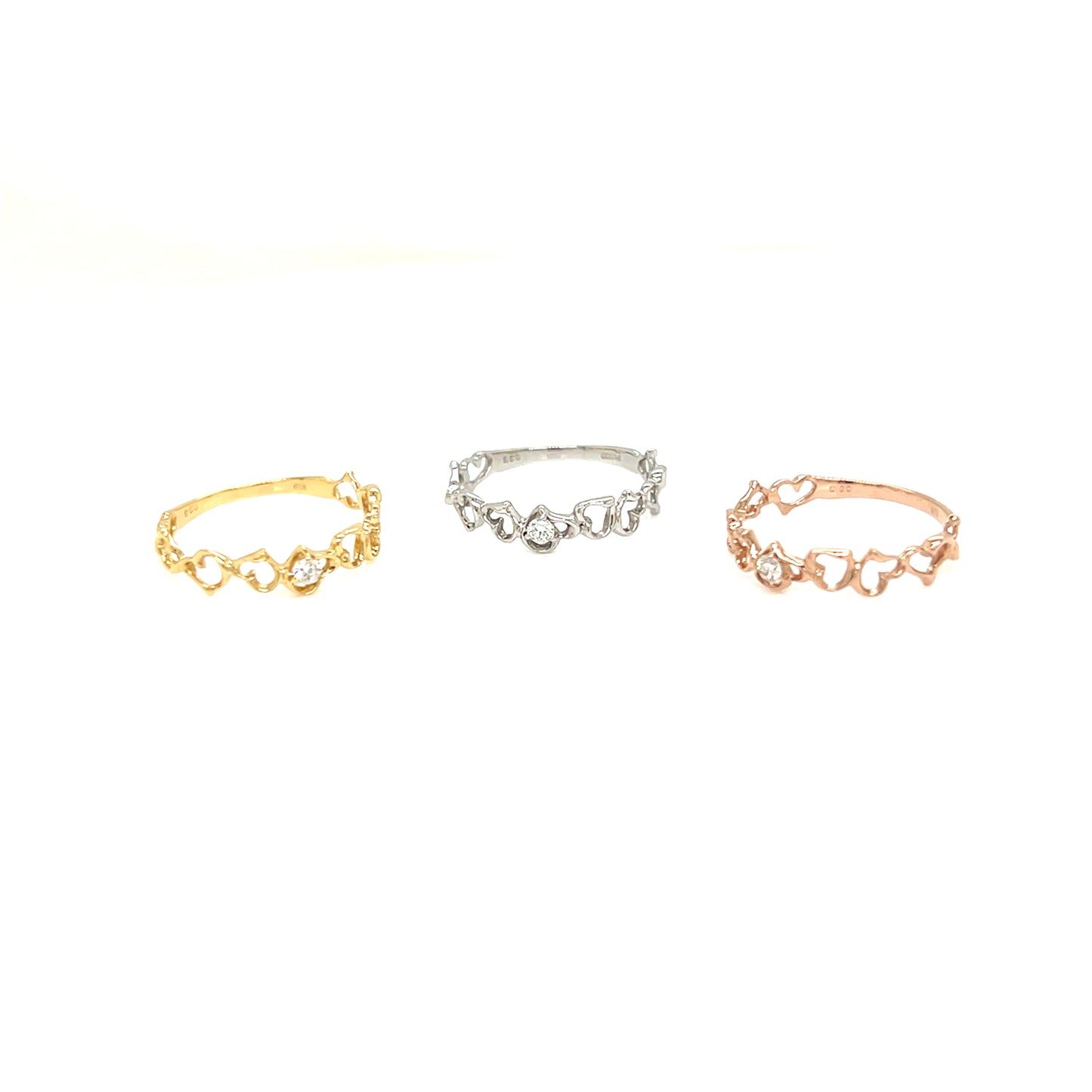 Three delicate rings with small heart-shaped links and clear stones, shown in gold, silver, and rose gold tones on a plain white background.