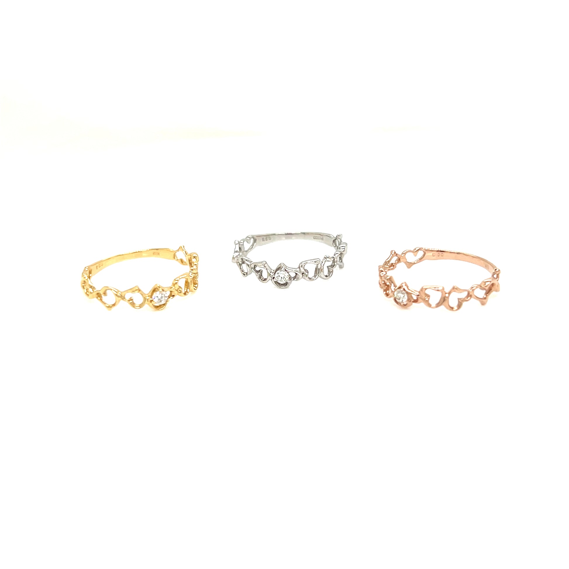 Three delicate rings with small heart-shaped links and clear stones, shown in gold, silver, and rose gold tones on a plain white background.
