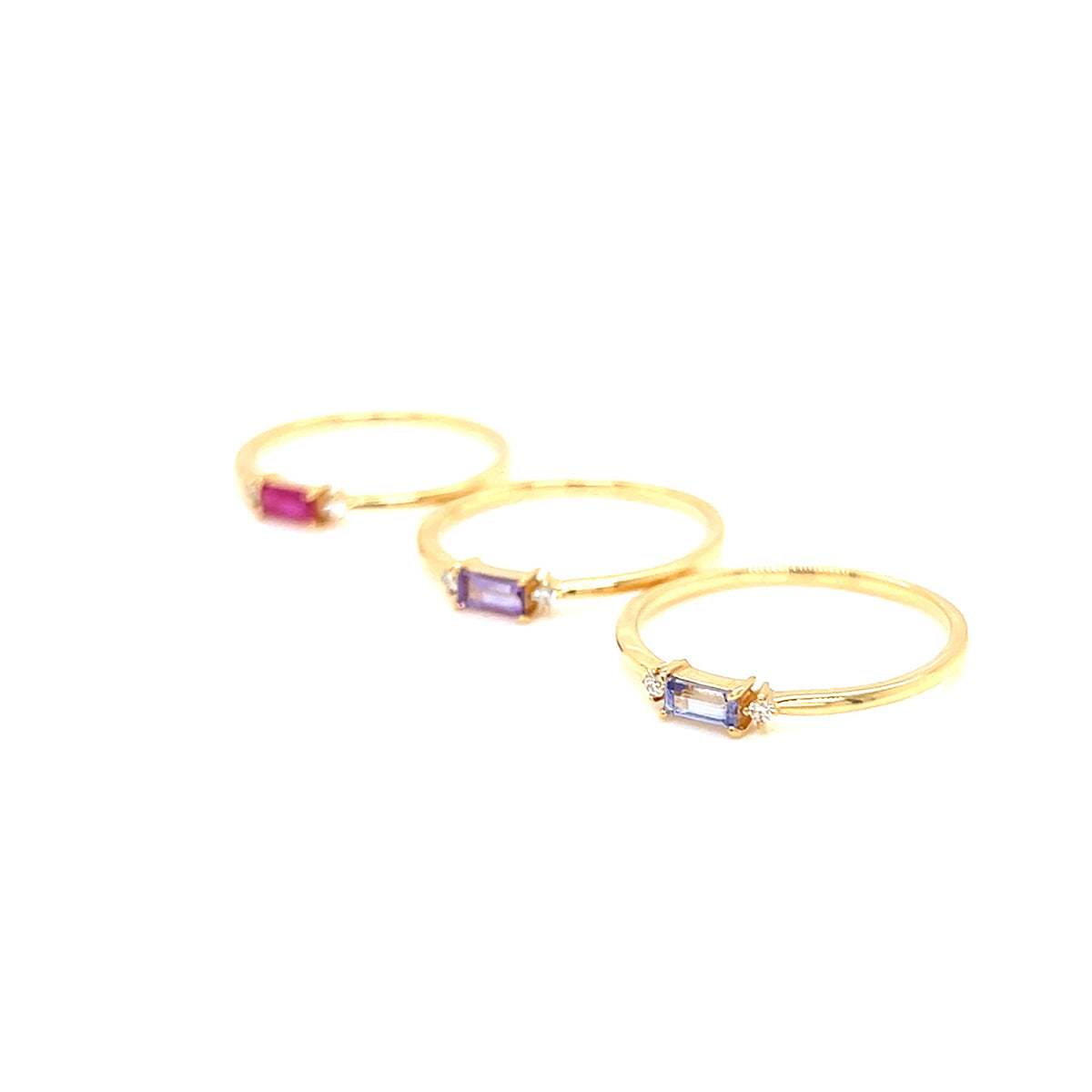 Three gold rings with small rectangular gemstones in pink, purple, and blue, arranged diagonally on a plain white background.