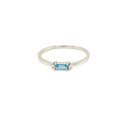 A delicate silver ring featuring a central light blue baguette-cut gemstone, photographed against a solid white background.