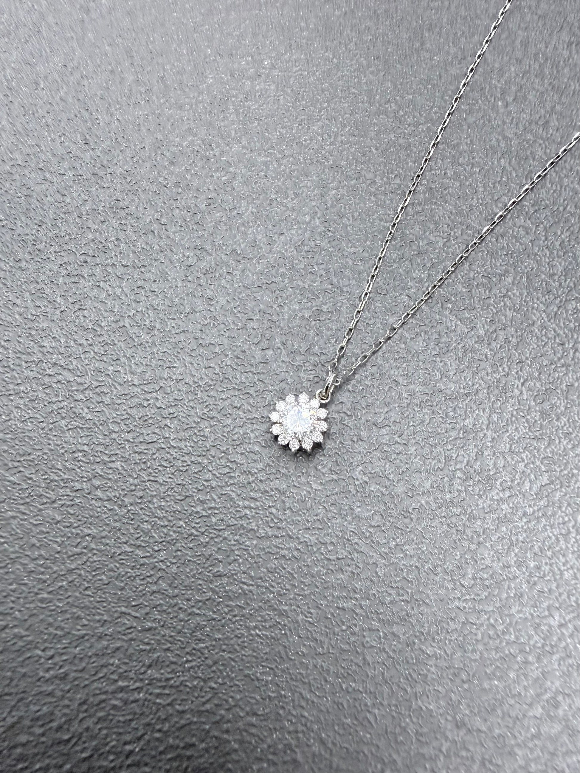A delicate silver chain necklace with a small flower-shaped pendant set with clear stones, displayed on a textured gray surface.