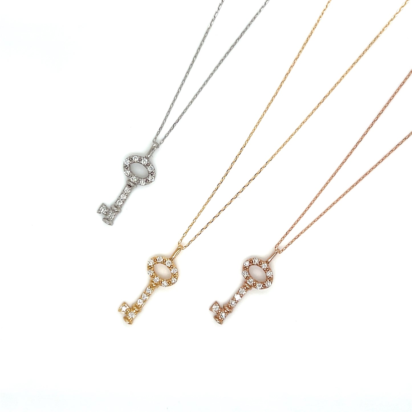 Three key-shaped pendant necklaces on delicate chains, shown in silver, yellow gold, and rose gold. Each pendant features a central oval white stone surrounded by small clear crystals.