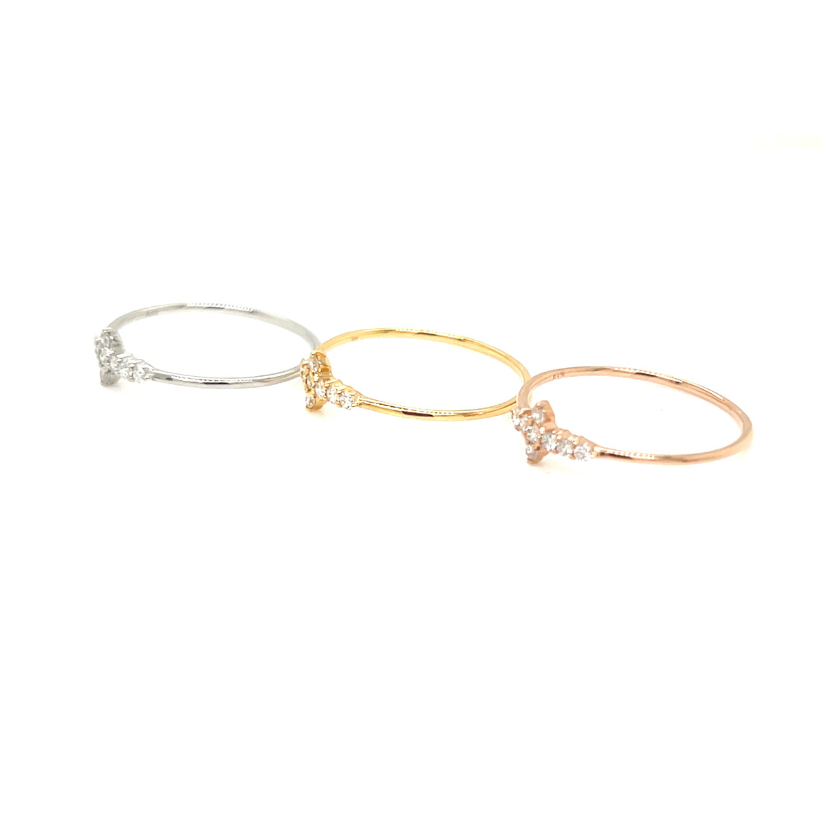 Three delicate bangle rings in silver, yellow gold, and rose gold, each featuring a small cluster of clear pavé stones at the center, arranged diagonally on a solid white background.