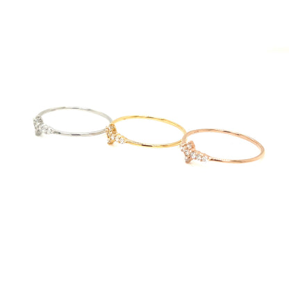 Three delicate bangle rings in silver, yellow gold, and rose gold, each featuring a small cluster of clear pavé stones at the center, arranged diagonally on a solid white background.