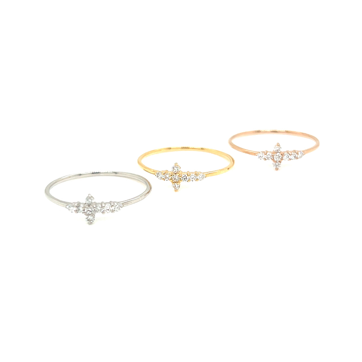 Three delicate cross-shaped rings in silver, yellow gold, and rose gold, each adorned with small clear gemstones, displayed on a clean white background.